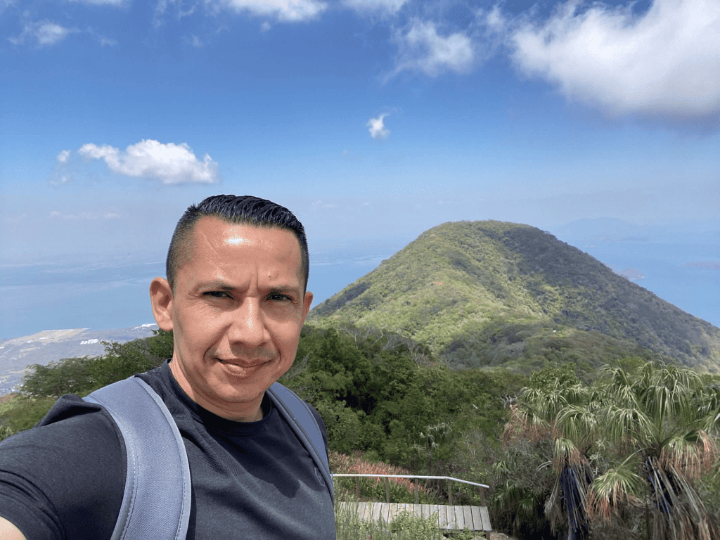 Mountain landscape with lush greenery and blue sky, featuring a man with a backpack in the foregroun.
